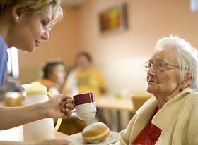 Ältere Frau bekommt Getränk und Essen von Pflegepersonal