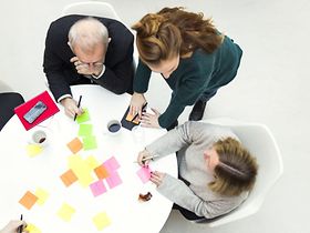 ältere und jüngere Mitarbeiter schreiben Klebezettel an Rundtisch