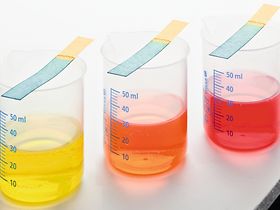 Drei Glasbecher mit farbigen Fluessigkeiten und Teststreifen