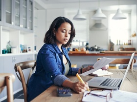 Shot of a young woman using a laptop and  going through paperwork while working from home