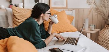 Frau liegt im Bett, trinkt Kaffee und ist an Laptop
