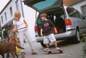 Frau mit Hund an der Leine und Kind auf Skateboard stehen vor geöffnetem Kofferraum