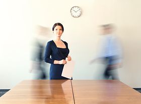 Frau steht im Büro an Konferenztisch, zwei Kollegen in Bewegung