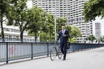 Geschäftsmann läuft mit Fahrrad an Brücke entlang und guckt auf sein smart phone