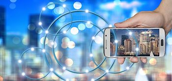 Hand mit Smartphone vor verschwommener Silhouette einer Großstadt