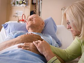 jüngere Frau sitzt am Bett eines alten Mannes und hält die Hand