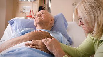 jüngere Frau sitzt am Bett eines alten Mannes und hält die Hand