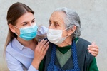 Portrait of friendly caregiver posing with elderly ill woman wearing surgical mask because of covid-