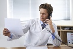 Shot of a concerned doctor sitting in his office and talking on the phone while looking over paperwo