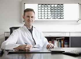 Junger Arzt sitzt am Schreibtisch und schaut in die Kamera