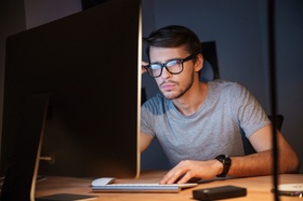 Junger Mann mit Brille sitzt vor Computer und schaut angestrengt auf Bildschirm