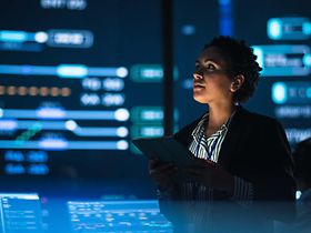 KI_Analystin mit Tablet vor Monitoring-Screens