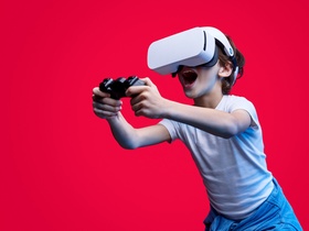 Casual boy wearing goggles of virtual reality while playing with gamepad on red backdrop