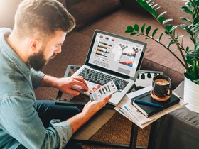 Young man, entrepreneur, freelancer sits at home on couch at coffee table, uses smartphone, working