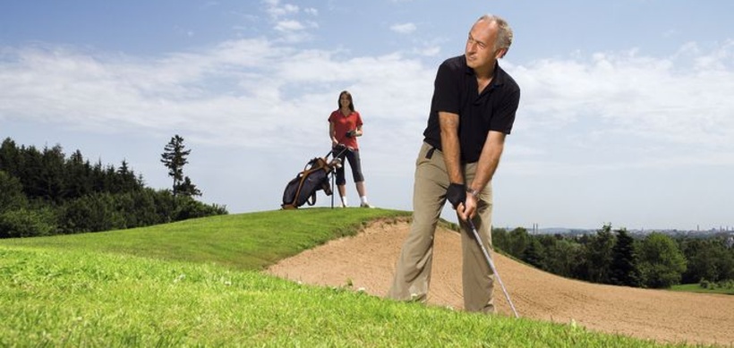 Aelterer Mann auf Golfplatz an Sandbunker, junge Fau mit Caddy im Hgr.