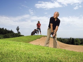 Aelterer Mann auf Golfplatz an Sandbunker, junge Fau mit Caddy im Hgr.