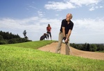 Aelterer Mann auf Golfplatz an Sandbunker, junge Fau mit Caddy im Hgr.