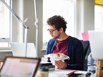 Mann isst Frühstück am Tisch vorm PC
