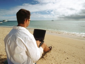 Mann mit Anzug und Laptop und sitzt am Starnd uns blickt aufs Meer