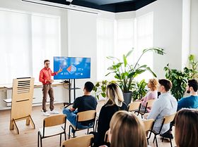 A seminar host using a large monitor as a visual aid and talking to the audience during a presentati