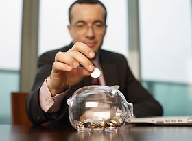 Mann wirft an Schreibtisch Muenze in Sparschwein