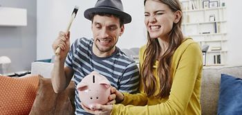 Paar sitzt auf Couch und schlägt mit Hammer auf Sparschwein