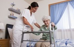 Pflegeperson hilft älterer Frau mit Gehhilfe vom Bett aufzustehen