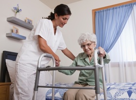 Pflegeperson hilft älterer Frau mit Gehhilfe vom Bett aufzustehen